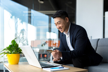 Asian boss transfers money from a bank card, sitting in the office, satisfied with the result businessman smiles, works on laptop