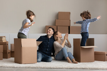 Happy little kid boy girl playing with big carton boxes, celebrating moving into own house with joyful couple parents. Bonding friendly family feeling excited renting new dwelling, real estate concept