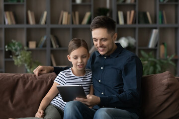 Happy bonding young father and little cute kid daughter using digital computer tablet applications, watching cartoons, shopping in internet store, playing online games, tech addiction concept.