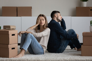 Unhappy exhausted young family couple sitting on floor carpet near carton boxes into new apartment,...