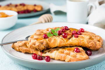 Crispy croissant waffles with red currants, honey and mint on plates and a cup of coffee on a light table. Sweet mouth-watering breakfast - croffles. Close-up