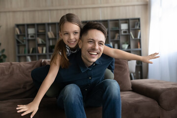 Fototapeta premium Happy young caring father holding on back adorable small kid daughter, playing airplane game sitting on cozy couch, entertaining involve in playtime weekend activity at home, single parenting concept.