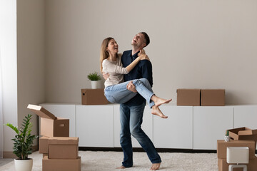 Affectionate young man holding in arms beautiful laughing wife, celebrating purchasing own dwelling. Happy millennial family couple having fun among big carton boxes in living room, relocation concept