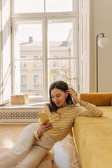 Obraz premium Attractive caucasian girl in headphones exchanging messages using application on her smartphone. Brunette is sitting on floor with her elbow on sofa in bright room. Telephone communication concept