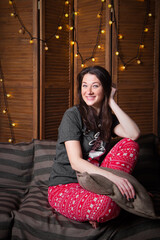 Fototapeta premium Beautiful romantic brunette woman in pajamas in cozy loft style room with garlands and rustic wooden screen. Portrait authentic female