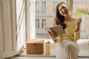 Happy young student is sitting on windowsill with workbook and enjoying excellent grade. Brown-haired woman clenches fist with her eyes closed, dressed in casual summer clothes. Exam passing concept