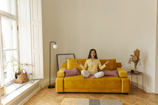 Portrait of young european woman sitting on sofa cross-legged and meditating with closed eyes. Brunette in concentrated state relaxes at home alone. Leisure lifestyle and beauty concept.