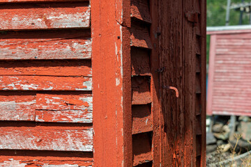 The exterior corner view of a vintage bright red outhouse bathroom or privy. The old wooden outdoor toilet has wood siding on the walls, a vintage handle or latch, and a single closed red wood door. 
