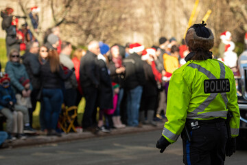 Police officer, wearing fluorescent clothing, standing in a crowded street. The officer is wearing a bulletproof vest with the word police on her back. A group of young people is in the background. 