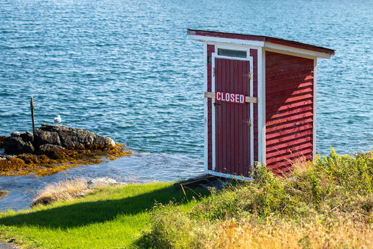 A small red building, outhouse, on the edge of a rocky cliff with blue ocean and islands in the background.  The outhouse has a closed sign across its door. The sky is blue with some white clouds. 