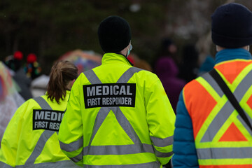 Medical first responders walking along a road wearing black wool stocking caps, yellow reflective coats with the medical first responder in grey letters and across. The EMT is carrying a first aid kit