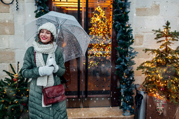 Happy woman walking on city street by decorated for Christmas festive restaurant entrance holding umbrella under snow