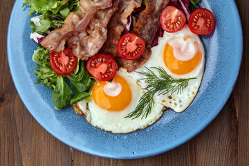 Fried eggs on a blue plate with bacon, lettuce and cherry tomatoes