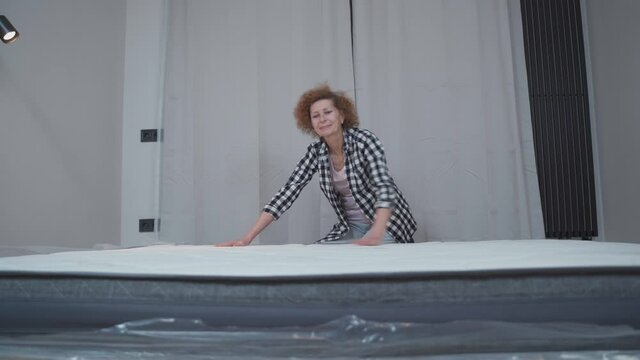 Elderly woman pleased with purchase new mattress in new rented apartment in empty bedroom. Mature female happy tries and inspects new sleeping mattress at home only after having moved to new home.