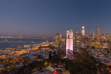 Nighttime aerial view of the San Francisco skyline with Coit Tower prominent in the frame. Bay Bridge in the background.