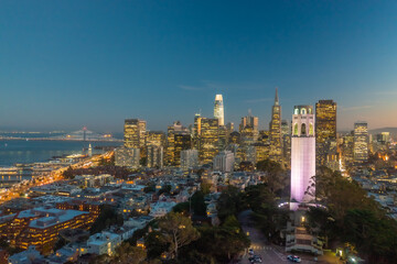 Nighttime aerial view of the San Francisco skyline with Coit Tower prominent in the frame. Bay Bridge in the background.