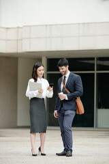 Positive young businesswoman reading notification on smartphone when talking to colleague outdoors