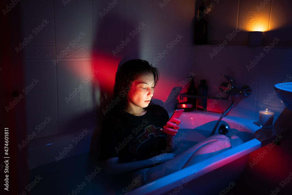 Wall mural young woman with smartphone sitting in bathtub full of water in dark bathroom with neon light and bu