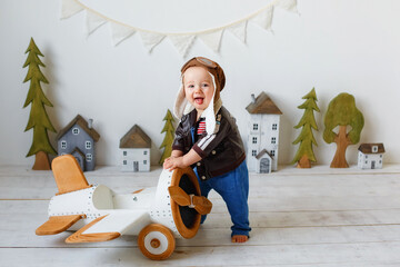 A happy one-year-old boy in a helmet and a pilot's jacket stands near a wooden plane. Portrait in...