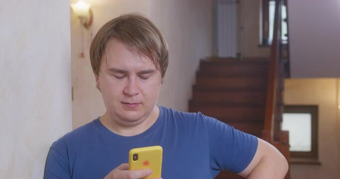 Concentrated Young Plump Man Types Message On Mobile Phone In Yellow Case Standing Against Big Wooden Stairs AT Home Close View