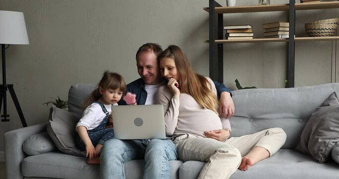 Happy young parents of little daughter expecting for second baby spend leisure time on sofa hug watch family photos funny cartoon on laptop. Smiling mother to be father toddler girl do shopping online