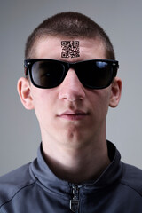 Young man with glasses presents a tattoo with a QR code on his forehead.