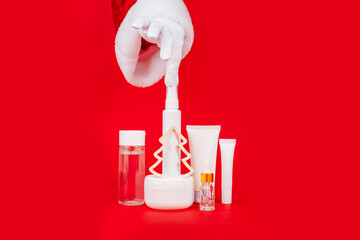 Santa's hand touches day cream cosmetic oil, eye cream on red background. Unbranded bottles in...