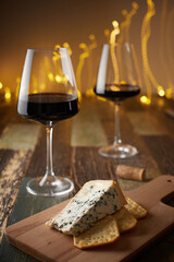 Still life of two glasses of wine, cheese with blue mold and crackers on a wooden board against the background of a New Year's garland. Front view.