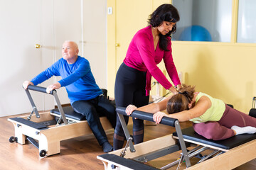 Fototapeta premium Hispanic female pilates instructor helping young woman doing exercises on reformer apparatus at gym..