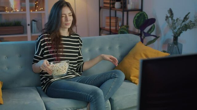 Slow motion of female student turning on TV and watching film eating popcorn at night in apartment. Youth lifestyle and leisure time activities concept.
