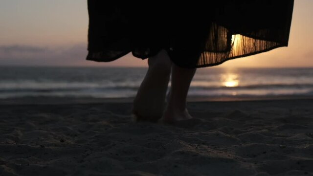 Close up of female legs in maxi black skirt, barefoot walking on the sand to the ocean, changing to panorama on the sunshine scape reflecting on the water 