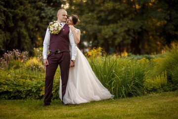 Bride and groom stand in a beautiful green garden on a warm autumn day. Newlyweds walk along the trees alley in the park. Wedding walk of the bride and groom at sunset.