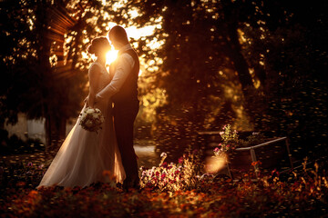 Bride and groom stand in a beautiful green garden on a warm autumn day. Newlyweds walk along the trees alley in the park. Wedding walk of the bride and groom at sunset.