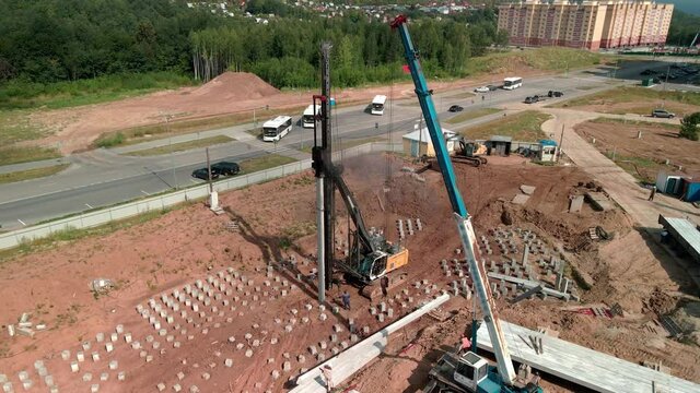 A large vertical pile-driving crane hammers reinforced concrete piles into the ground at a construction site.