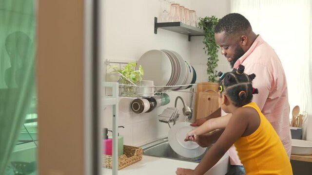 4K African father teaching little daughter washing dishes in the sink on kitchen counter with talking together after having dinner. Happy family dad and child girl kid spending time together at home.