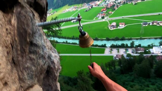 POV shot of a climber who rings a bell, which is located at very high altitude. You can only see his right muscular arm. Beneath him is a valley with trees, a beautiful blue river and a green meadow