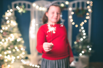 Beautiful little girl sitting near nicely decorated Christmas tree, holding sparklers and having fun while celebrating New Year's Eve