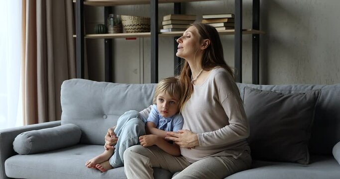 Young pregnant female sit on couch at living room cuddle son preschooler with love care. Loving expectant mom caress elder child hold hand of little boy on big belly to let feel kicks of baby inside