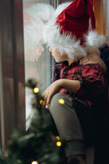 cute caucasian boy wearing santa hat and plaid shirt sitting by the window waitinf for christmas