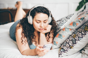 Overweight young woman with loose hair in top and shorts listens to music with wireless headphones holding smartphone sitting on double bed