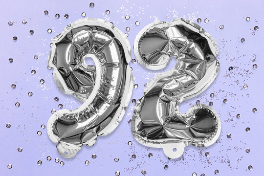 Silver foil balloon number, digit ninety two on a lilac background with sequins. Birthday greeting card with inscription 92. Top view. Numerical digit. Celebration event, template.