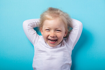 Naklejka premium Smiling beautiful blonde girl lying down holds her hands behind her head on a blue background. Top view, flat lay.