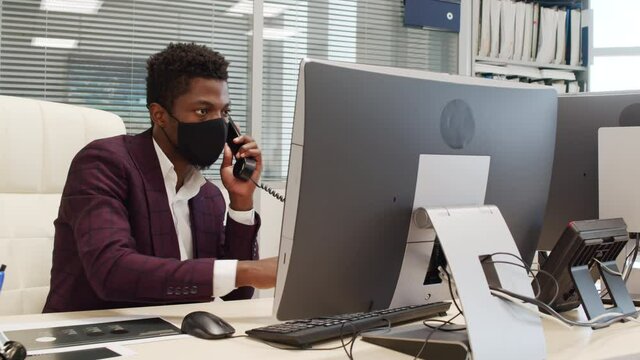 Handheld medium shot of African-American manager in suit and face mask sitting at desk in office and talking on phone while typing on computer