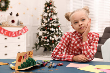 Cute little child with materials and beautiful Christmas card at table in decorated room