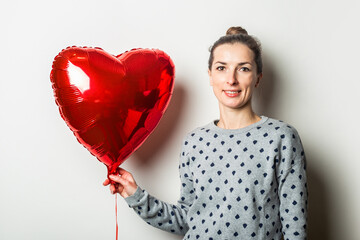 Smiling young woman in a sweater holding a heart air balloon on a light background. Valentine's Day...