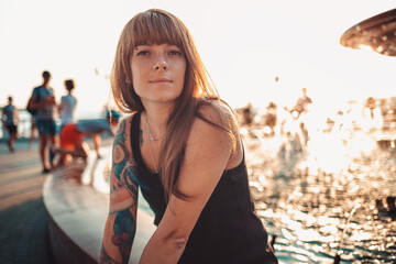 Portrait of tanned young woman with a tattoo on her arm is sitting by the fountain. Sunset in the background