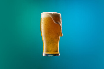 Full glass of frothy light lager beer isolated over gradient blue and green color background in...