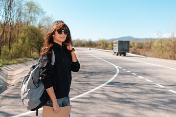 Fototapeta premium Hitchhiking. Smiling young woman in sunglasses and cap holding a cardboard sign. Country road on the background. Copy space. The concept of local traveling