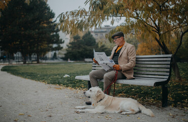 Happy senior man sitting on bench and reading newspaper during dog walk outdoors in park in city.