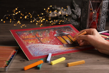 Woman drawing beautiful lantern with soft pastel at wooden table against blurred lights, closeup